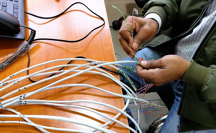 A person is working on a bundle of network cables, stripping and twisting the wires together. The cables are laid out on a wooden desk, and the person is using their hands to manage the wires. A phone and other equipment are visible on the desk.