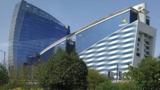 "Two modern, uniquely designed buildings with blue glass facades and white wave-like patterns, surrounded by greenery. One building has a logo with a yellow triangle and the text 'DLF' on it. The buildings are connected by a skybridge.