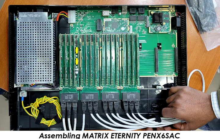 A person is assembling a MATRIX ETERNITY PENX6SAC device. The image shows the internal components of the device, including multiple green circuit boards with various electronic components and connectors. Several white cables are connected to the bottom of the circuit boards. The person's hand is visible, holding one of the cables. The text 'Assembling MATRIX ETERNITY PENX6SAC' is displayed at the bottom of the image.