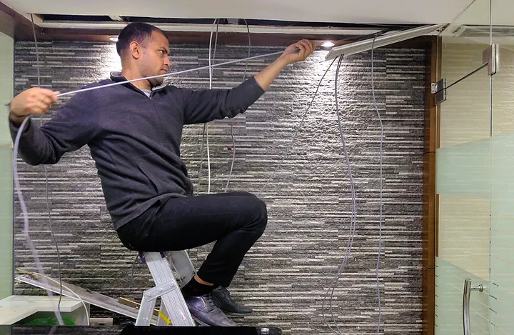 A person is sitting on a step ladder, working on electrical wiring in a room. The ceiling panels are removed, exposing the wires. The person is holding and managing several cables, appearing to be in the process of installation or repair. The background features a textured stone wall and a glass door.