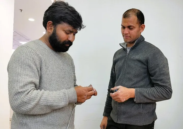 "Two people standing indoors, both holding small electronic devices. One person is wearing a light gray sweater, and the other is wearing a dark gray jacket.