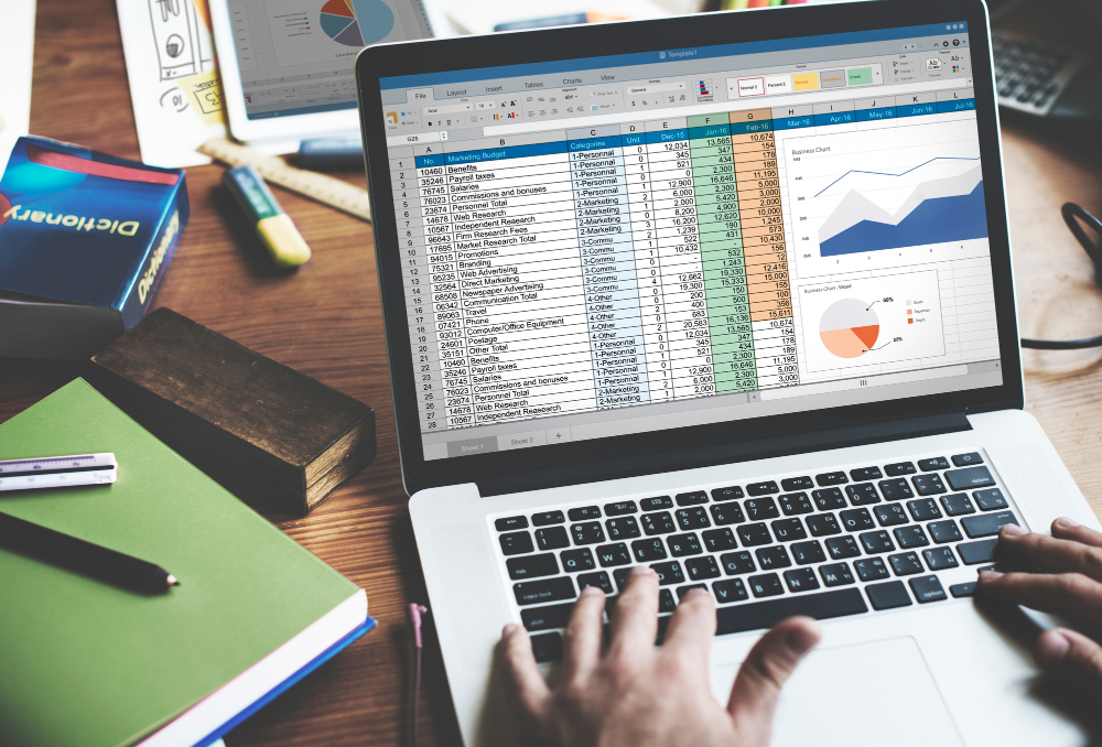 A person is working on a laptop displaying a spreadsheet with a marketing budget. The spreadsheet includes various columns such as "ID," "Description," "Category," "CWC," and monthly expenses from January to June.