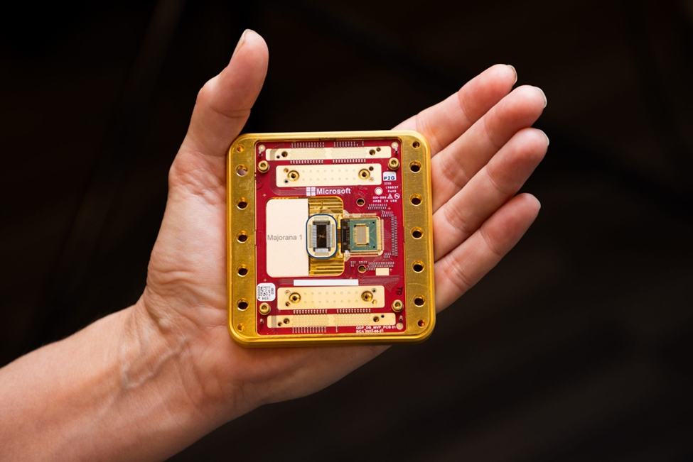 A hand is holding a square-shaped electronic component with a red and gold circuit board. The circuit board has various components, including a central chip labeled "Microsoft" and "Majorana 1." The board also features multiple connectors and gold-plated elements. The background is dark, making the circuit board stand out prominently