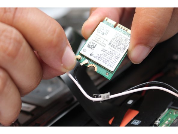 A close-up image of a person's hands installing or removing a wireless network card from a laptop. The card has various technical details and QR codes printed on it. Two wires, one black and one white, are being connected to the card.